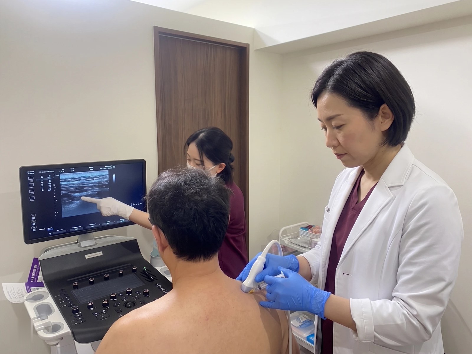 医師がエコーで確認しながら肩に筋膜リリース注射を行い、スタッフがモニターで説明している様子